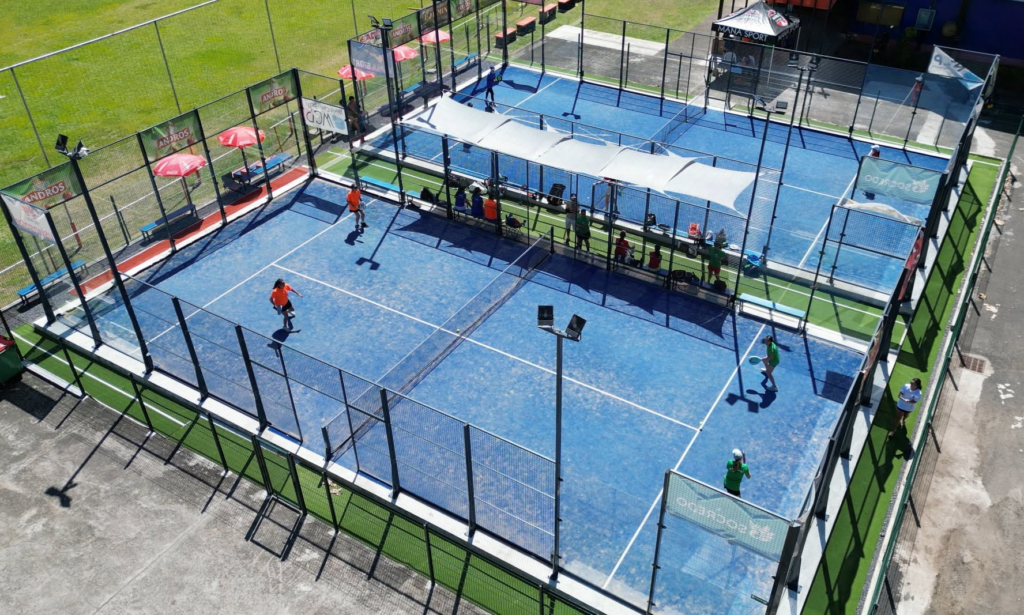 Aerial view of blue padel courts with players in orange and green shirts, surrounded by tall fences and benches along the sidelines.