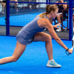Female padel player in a blue outfit lunges to strike a ball with a perforated paddle on a bright blue court, behind a blue fence with spectators.