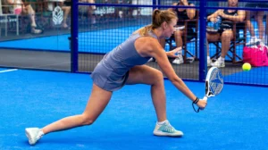 Female padel player in a blue outfit lunges to strike a ball with a perforated paddle on a bright blue court, behind a blue fence with spectators.