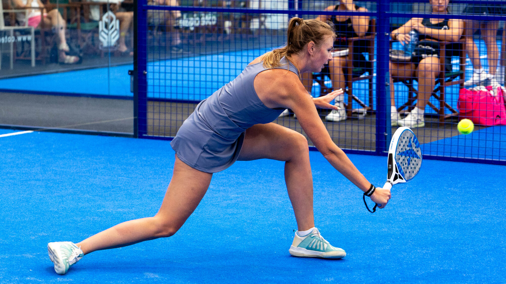 Female padel player in a blue outfit lunges to strike a ball with a perforated paddle on a bright blue court, behind a blue fence with spectators.