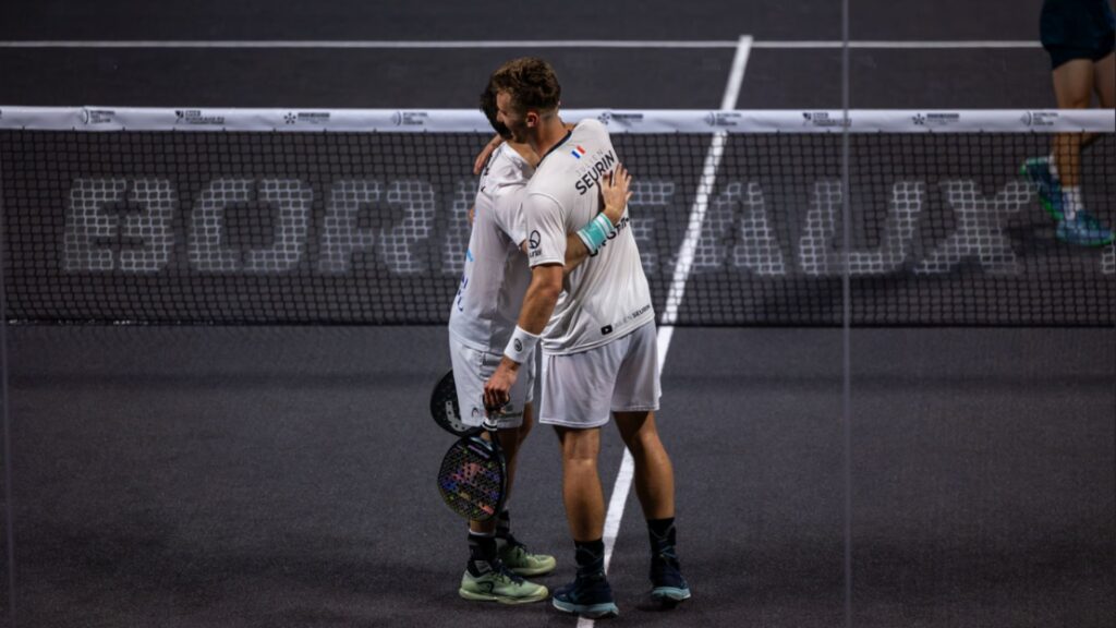 Two tennis players in white uniforms hug at the net after a match, on a dark court with a visible boundary line kept between them.