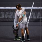 Two tennis players in white uniforms hug at the net after a match, on a dark court with a visible boundary line kept between them.