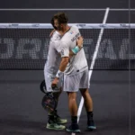 Two tennis players in white uniforms hug at the net after a match, on a dark court with a visible boundary line kept between them.