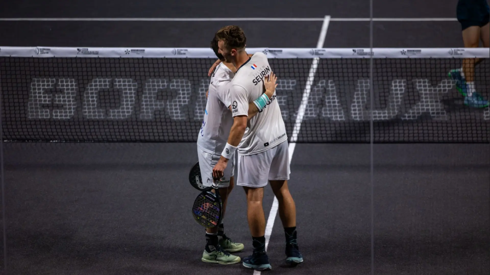 Two tennis players in white uniforms hug at the net after a match, on a dark court with a visible boundary line kept between them.