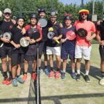 Group of players posing for a photo on an outdoor court, each holding a paddle and wearing helmets and athletic clothing, with a net visible in the foreground.
