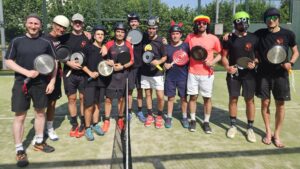 Group of players posing for a photo on an outdoor court, each holding a paddle and wearing helmets and athletic clothing, with a net visible in the foreground.