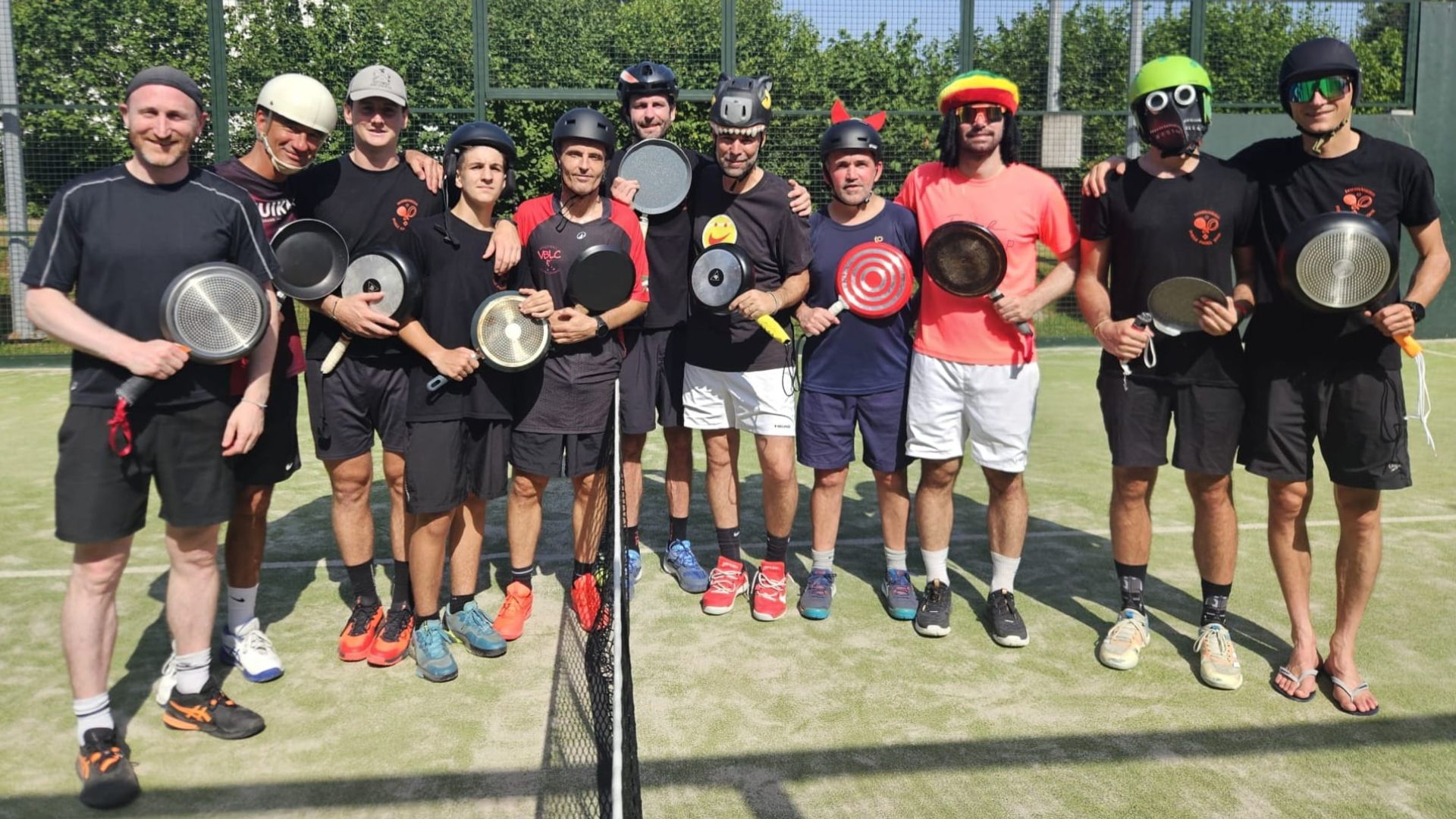 Group of players posing for a photo on an outdoor court, each holding a paddle and wearing helmets and athletic clothing, with a net visible in the foreground.