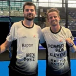 Two smiling padel players in matching white jerseys posing with a paddle on an indoor blue court after a match.