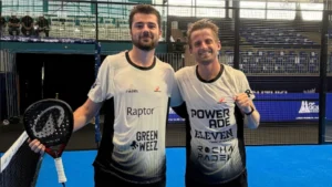 Two smiling padel players in matching white jerseys posing with a paddle on an indoor blue court after a match.