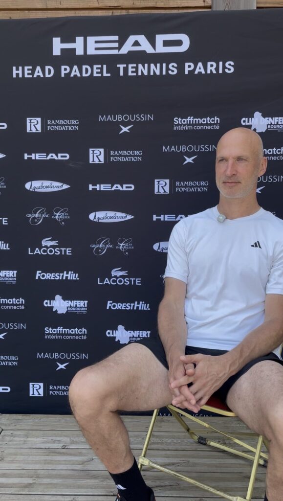 Man in a white athletic shirt sits on a chair in front of a black sponsor backdrop at an event.