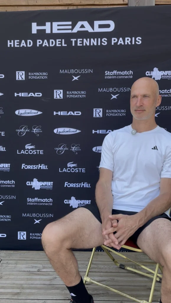 Screenshot | Padel Magazine Man in a white athletic shirt sits on a chair in front of a black sponsor backdrop at an event.