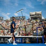 Two female padel players on a blue court celebrate a point as a large crowd watches from the stands outside under a sunny sky.