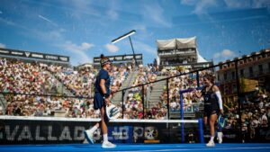 Two female padel players on a blue court celebrate a point as a large crowd watches from the stands outside under a sunny sky.