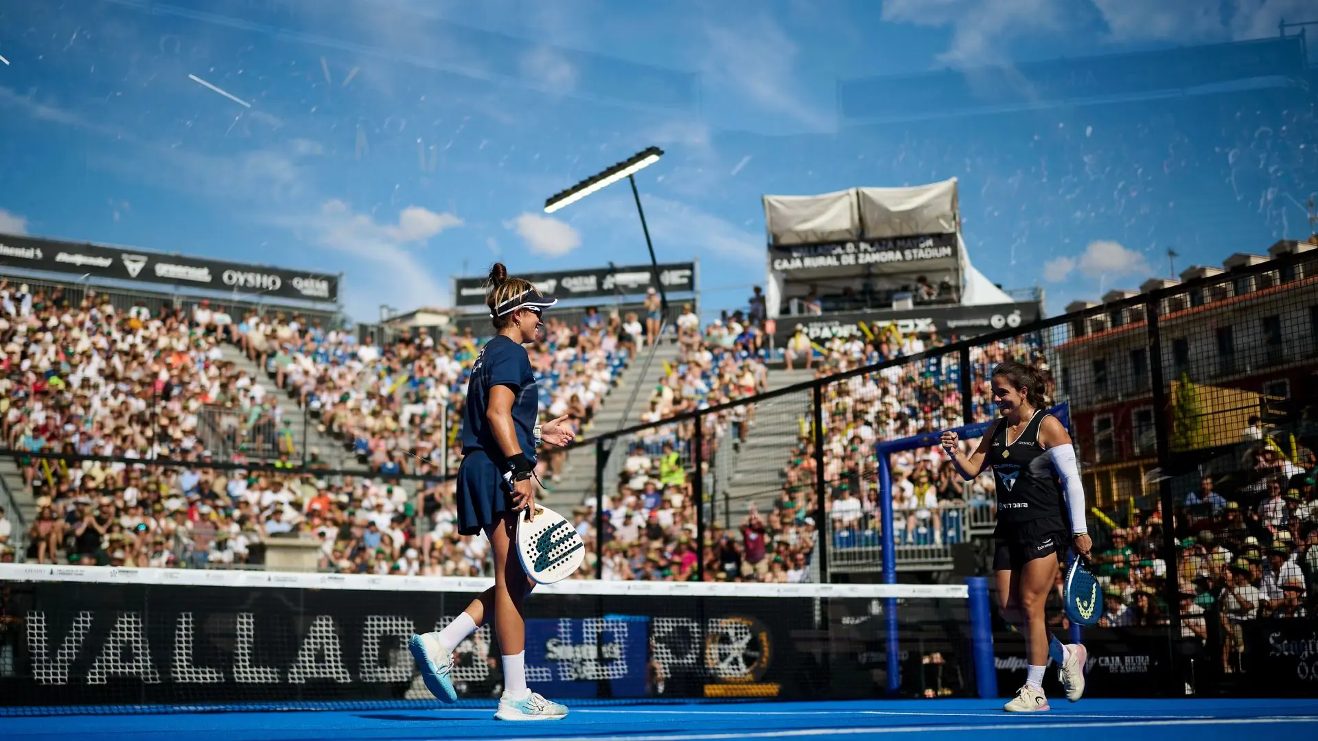 Two female padel players on a blue court celebrate a point as a large crowd watches from the stands outside under a sunny sky.