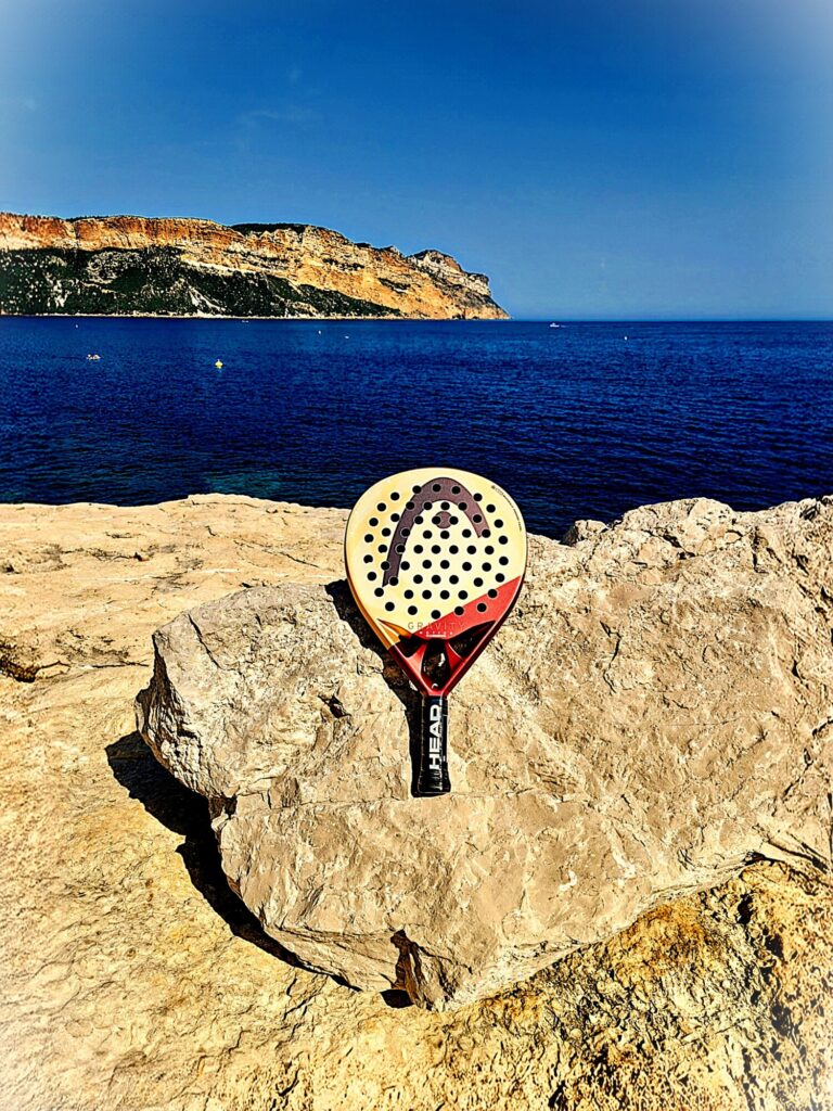 Padel racket wedged into a rocky ledge along a calm blue sea with cliffs in the background.