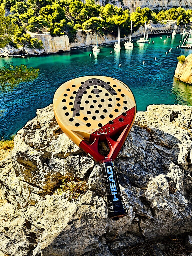 Close-up of a large perforated circular disk resting on rocky edge, with turquoise harbor and anchored boats in the background. The disk has holes and a red handle/attachment at the bottom.