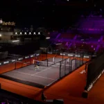 Indoor badminton arena with multiple courts, players on the foreground court, and spectators in red seats under purple lighting