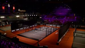 Indoor badminton arena with multiple courts, players on the foreground court, and spectators in red seats under purple lighting
