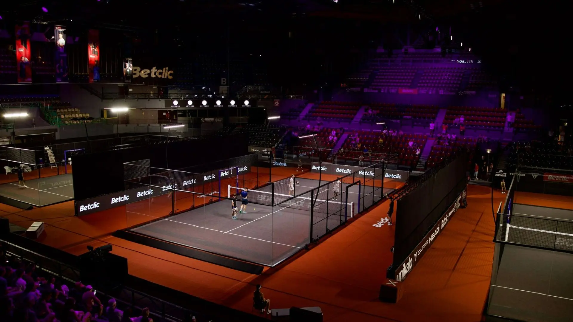 Indoor badminton arena with multiple courts, players on the foreground court, and spectators in red seats under purple lighting
