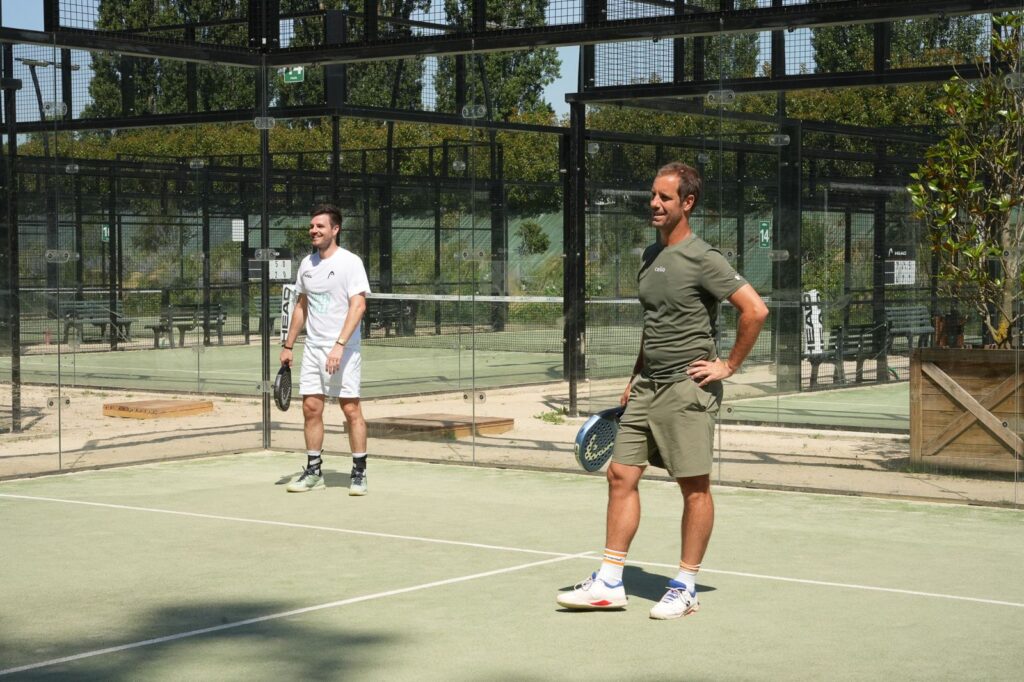 Two men stand on a padel court with rackets, ready for play, standing near a glass enclosure on a sunny day