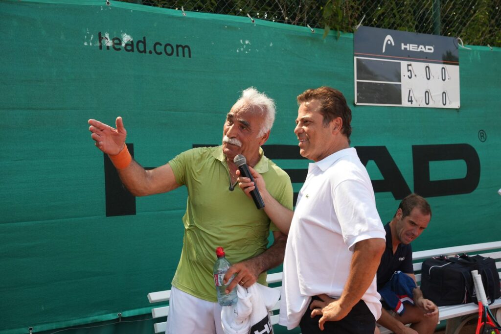 Two men on a tennis court backdrop; one in lime shirt with a microphone explaining something, the other in white smiling as he listens, a water bottle and towel in hand.