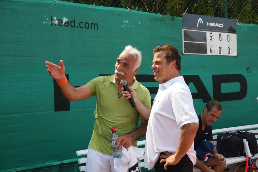Immagine WhatsApp 2026-06-29 alle 164155 | Padel Magazine Due uomini sullo sfondo di un campo da tennis; uno con una camicia verde lime e un microfono in mano che spiega qualcosa, l'altro in camicia bianca sorridente mentre ascolta, con una bottiglia d'acqua e un asciugamano in mano.