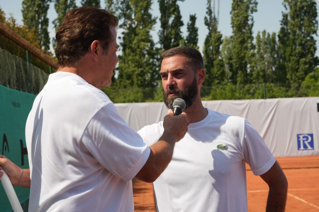 Two men in white shirts converse outdoors on a clay tennis court, one holding a microphone to the other as he speaks.