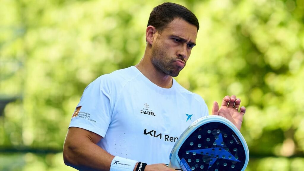 Man in a light blue shirt holds a padel racket outdoors, looking focused.