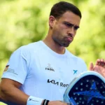 Man in a light blue shirt holds a padel racket outdoors, looking focused.