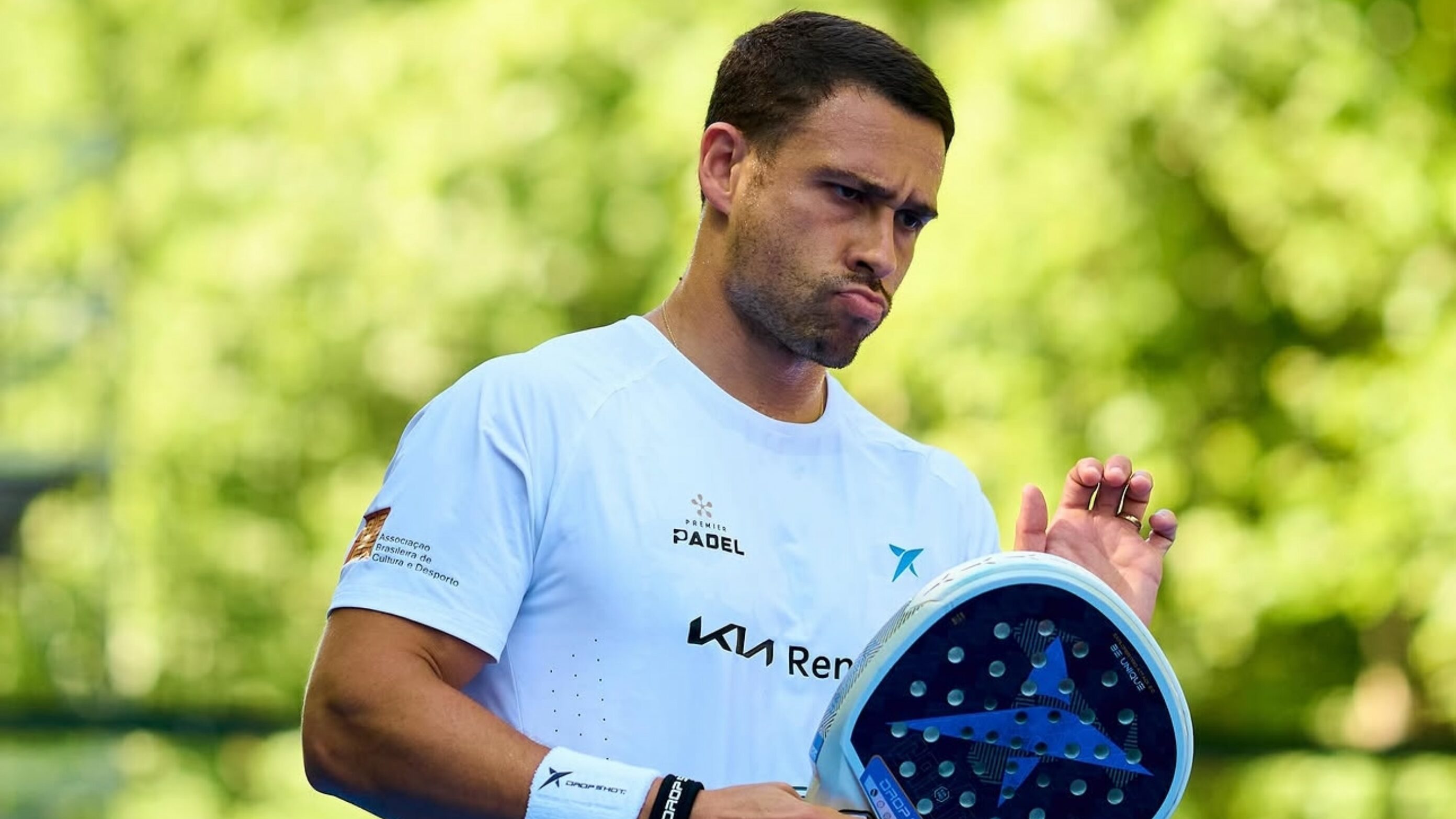 Man in a light blue shirt holds a padel racket outdoors, looking focused.