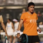Female athlete in an orange shirt holding a paddle-style racket on a court, with teammates blurred in the background.