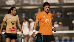 Female athlete in an orange shirt holding a paddle-style racket on a court, with teammates blurred in the background.