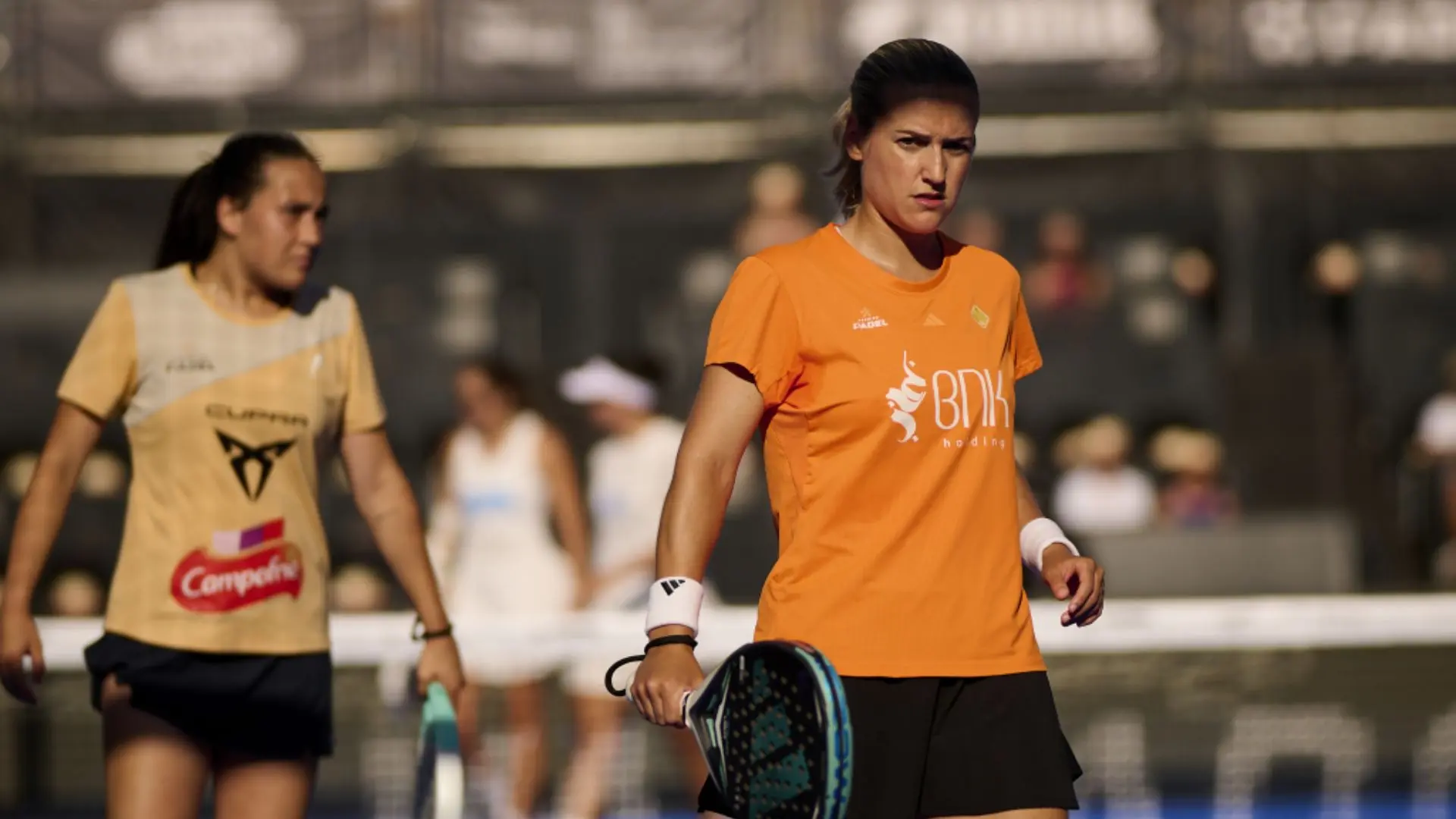 Female athlete in an orange shirt holding a paddle-style racket on a court, with teammates blurred in the background.