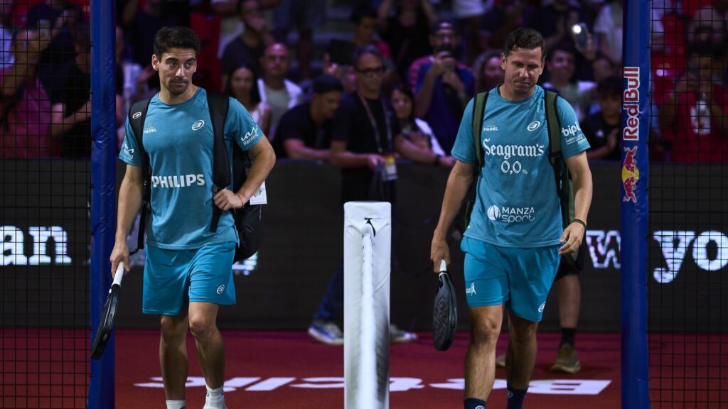 Two male padel players in turquoise outfits walk off a court with paddles as a crowd watches behind a net fence.