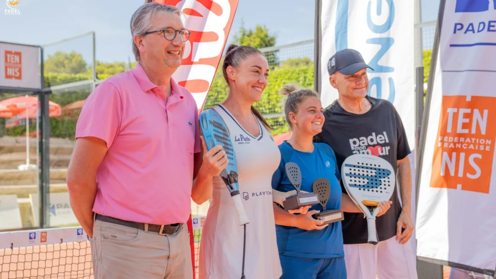 Cztery osoby pozują z paletkami do padla i trofeami na plenerowym wydarzeniu wręczania nagród, uśmiechając się obok banerów sponsorów.