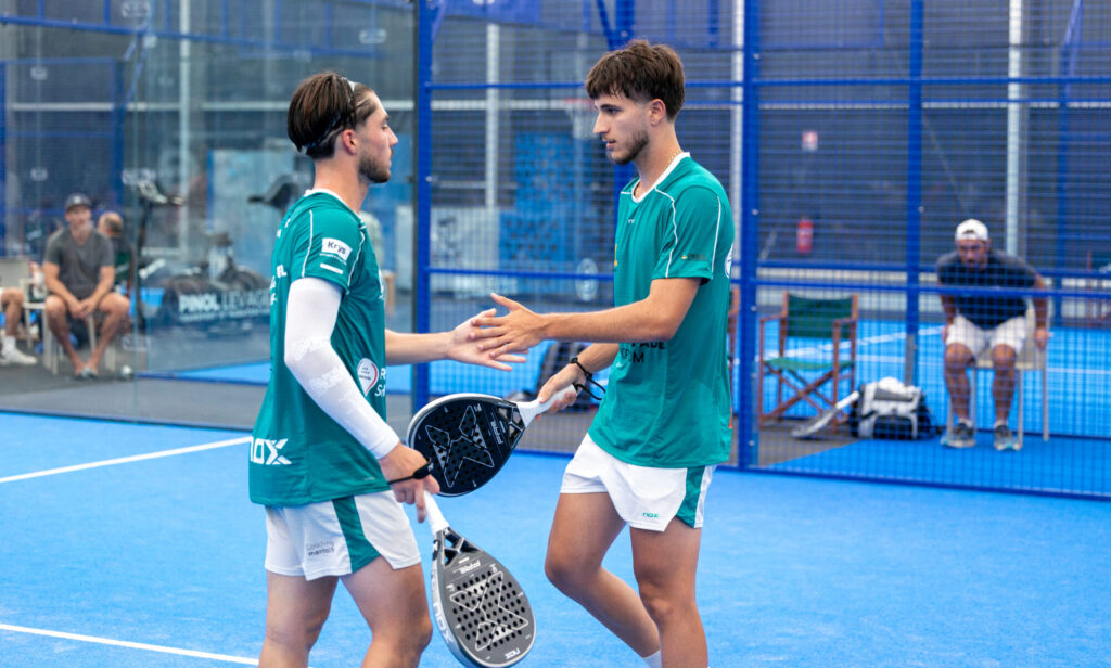 Two padel players in teal jerseys shake hands at the net on a blue indoor court, each holding a paddle.