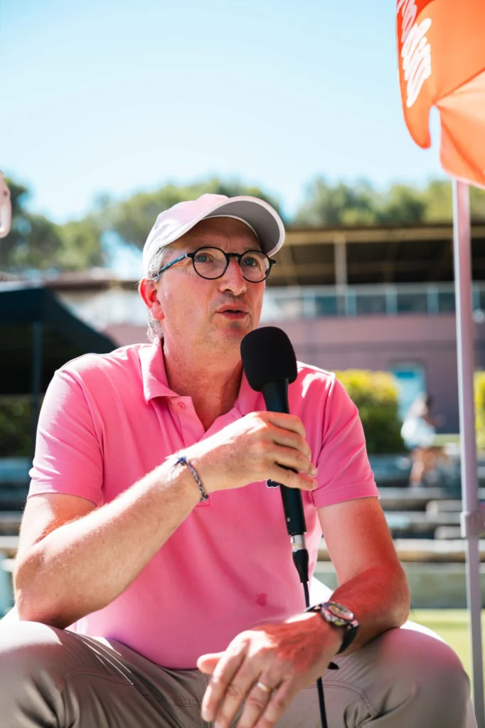 Man in a pink polo and white cap speaks into a handheld microphone outdoors at a sunny event.