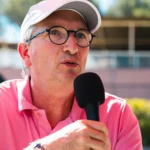 168A5198 - Copie | Padel Magazine Man in a pink polo and white cap speaks into a handheld microphone outdoors, wearing glasses.