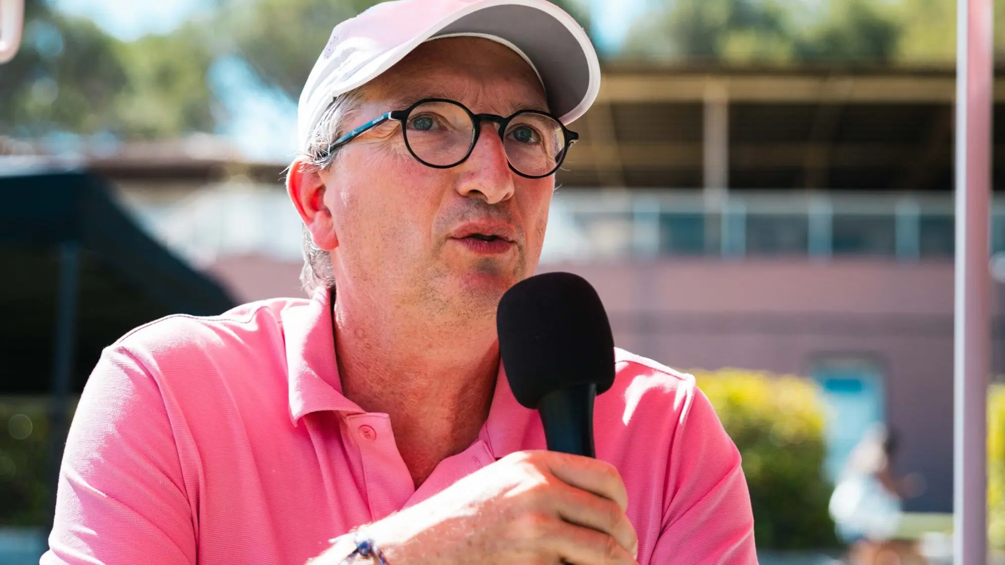 Man in a pink polo and white cap speaks into a handheld microphone outdoors, wearing glasses.