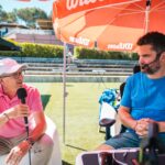 Two men interview outdoors on a sunny sports field under a large red umbrella; one in a pink polo speaks into a handheld mic, the other in a blue shirt wears a headset.