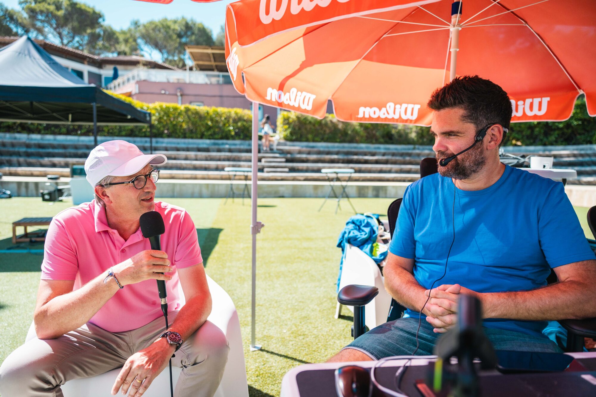 Two men interview outdoors on a sunny sports field under a large red umbrella; one in a pink polo speaks into a handheld mic, the other in a blue shirt wears a headset.