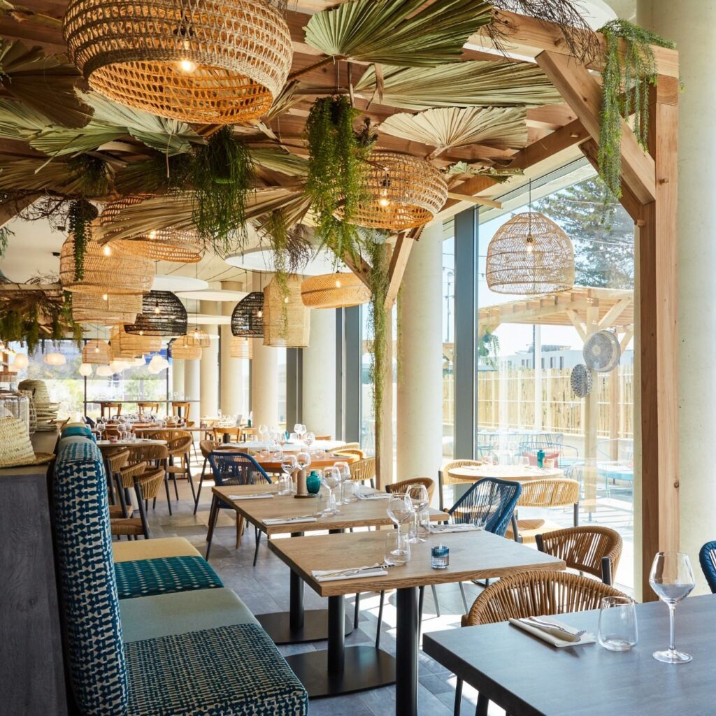 Restaurant interior with hanging wicker pendant lights, green plants, and wooden beams; tables set for dining by large windows.