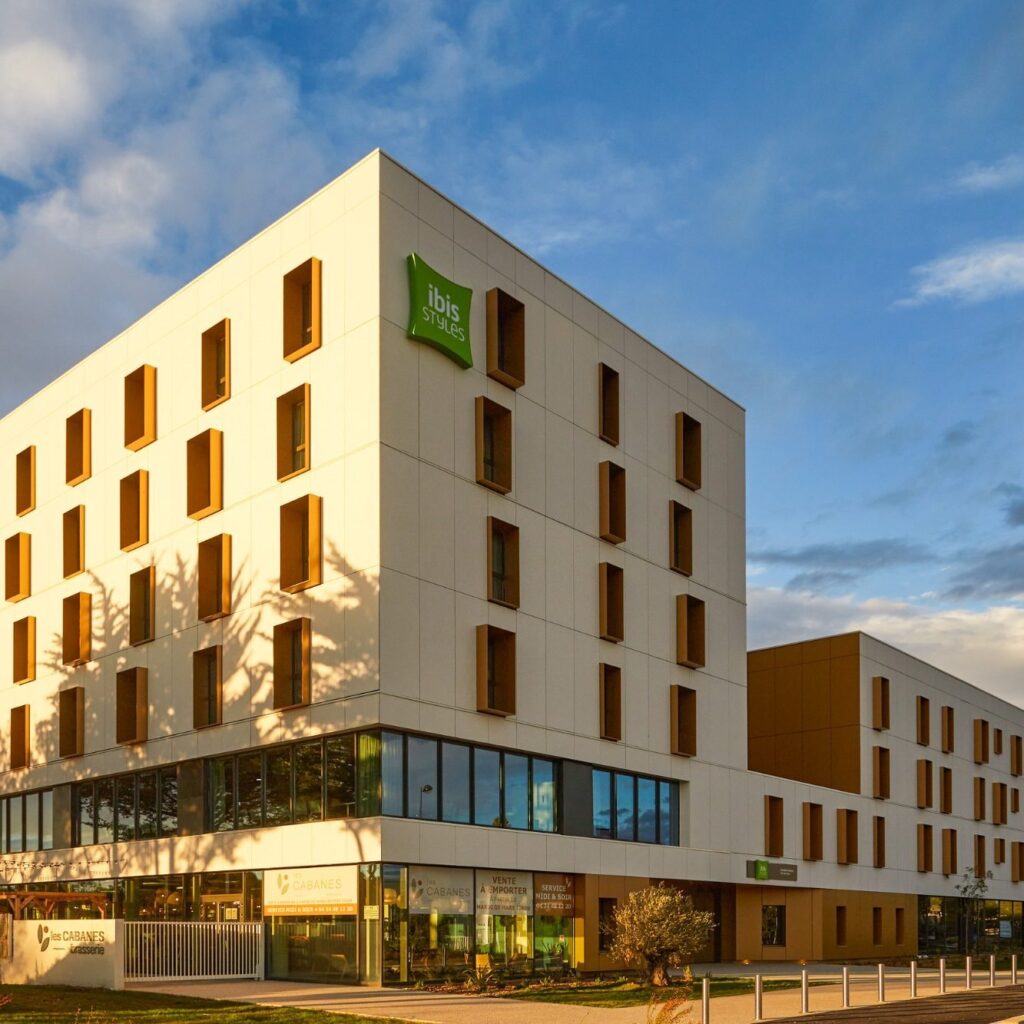 Modern white hotel building with a green ibis Styles sign and numerous rectangular windows against a blue sky.