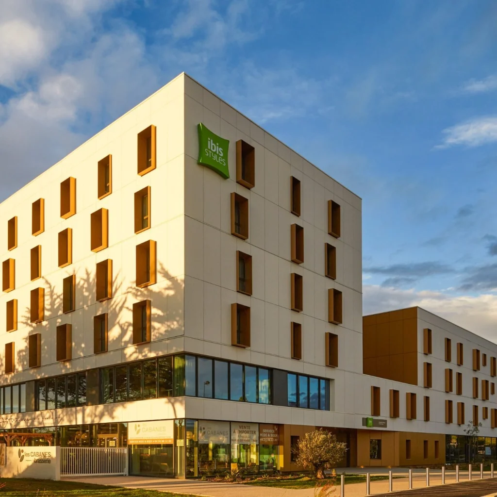 Moderni valkoinen hotellirakennus, jossa on vihreä ibis Styles -kyltti ja lukuisia suorakaiteen muotoisia ikkunoita sinistä taivasta vasten.