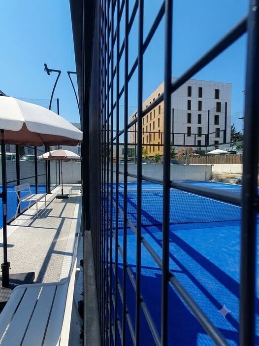 Pool area with blue-tiled water on the right, white umbrellas and benches along the left, and a metal fence grid in the foreground; a modern building rises in the background under a clear blue sky.
