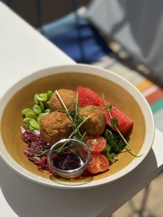Bowl of falafel with greens, edamame, cherry tomatoes, a watermelon cube, and a small cup of dipping sauce side-by-side.