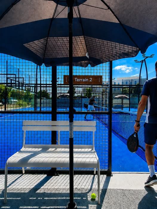 A white bench under a large umbrella in front of a blue-paved tennis or padel court, with a sign reading'Terrain 2' and a tennis ball on the ground nearby.