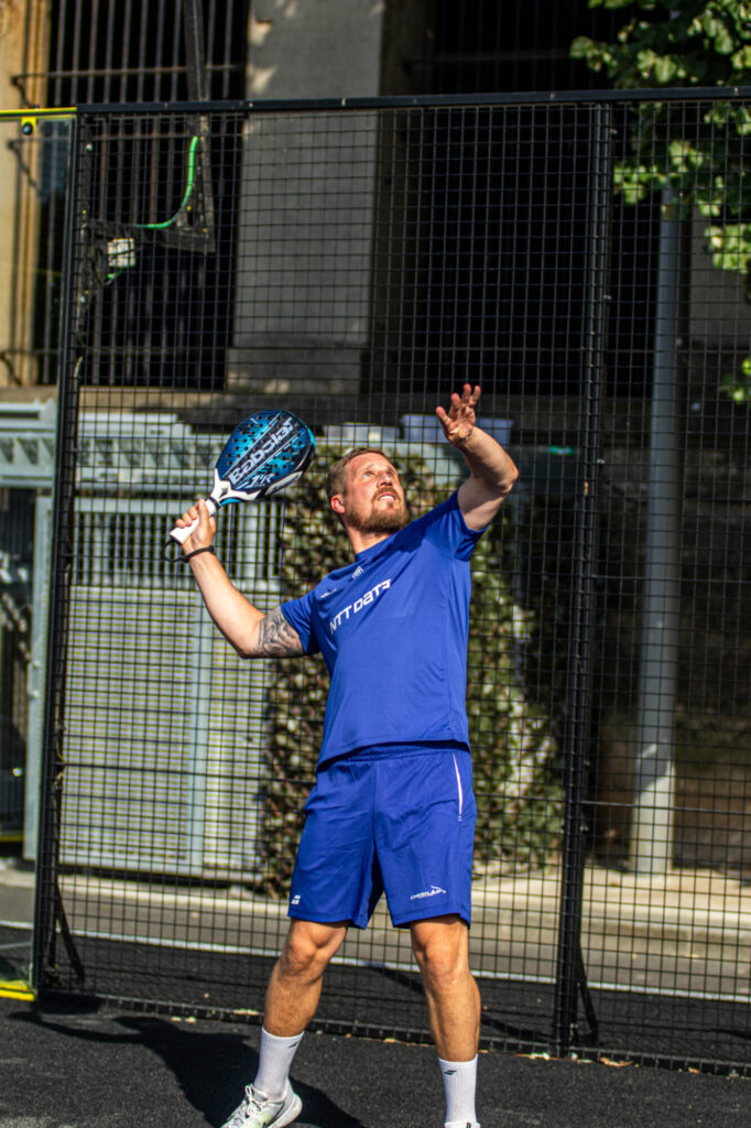 369A7568 | Padel Magazine Man in blue athletic gear holds a blue paddle racket on a padel court, ready to hit a ball, standing behind a tall black mesh fence.