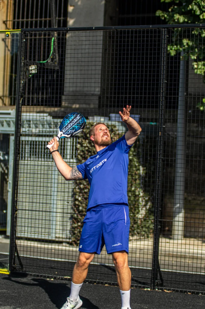 Man in blue athletic gear holds a blue paddle racket on a padel court, ready to hit a ball, standing behind a tall black mesh fence.