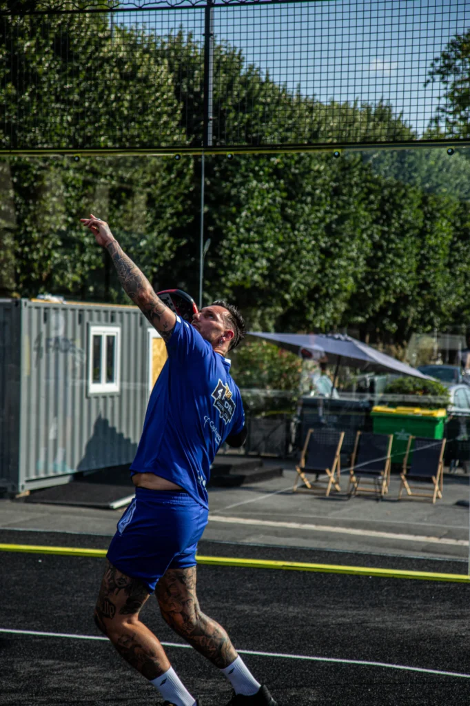 369A7696 | Padel Magazine Man in a blue sports shirt with tattoos on his arm reaching upward on an outdoor court under a high net, ready to hit a ball.